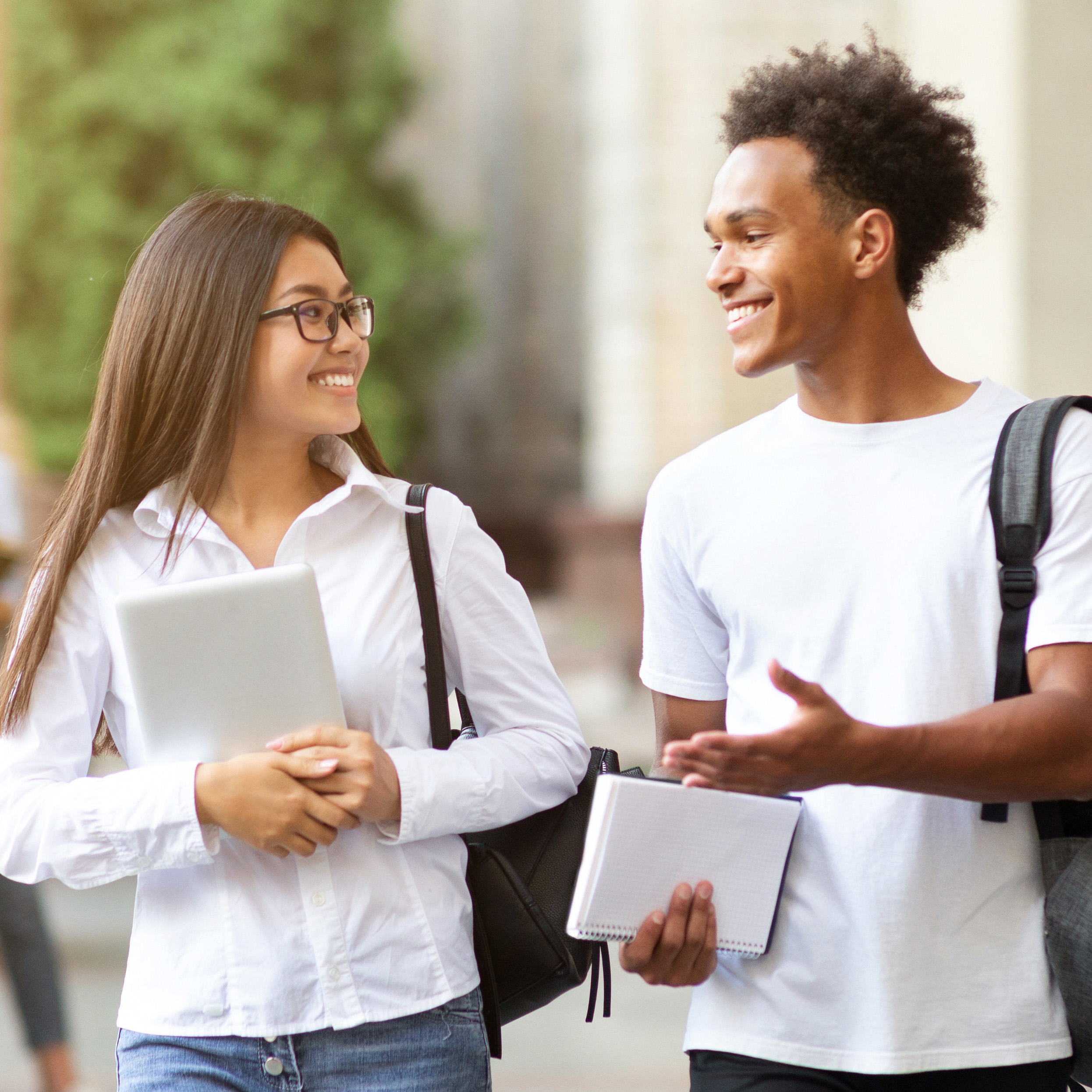 Eine junge Studentin und ein junger Student mit Schreiblöcken in der Hand und Tasche über der Schulter gehen redend über einen Universitätscampus. adobestock_281428326-1-1.jpg